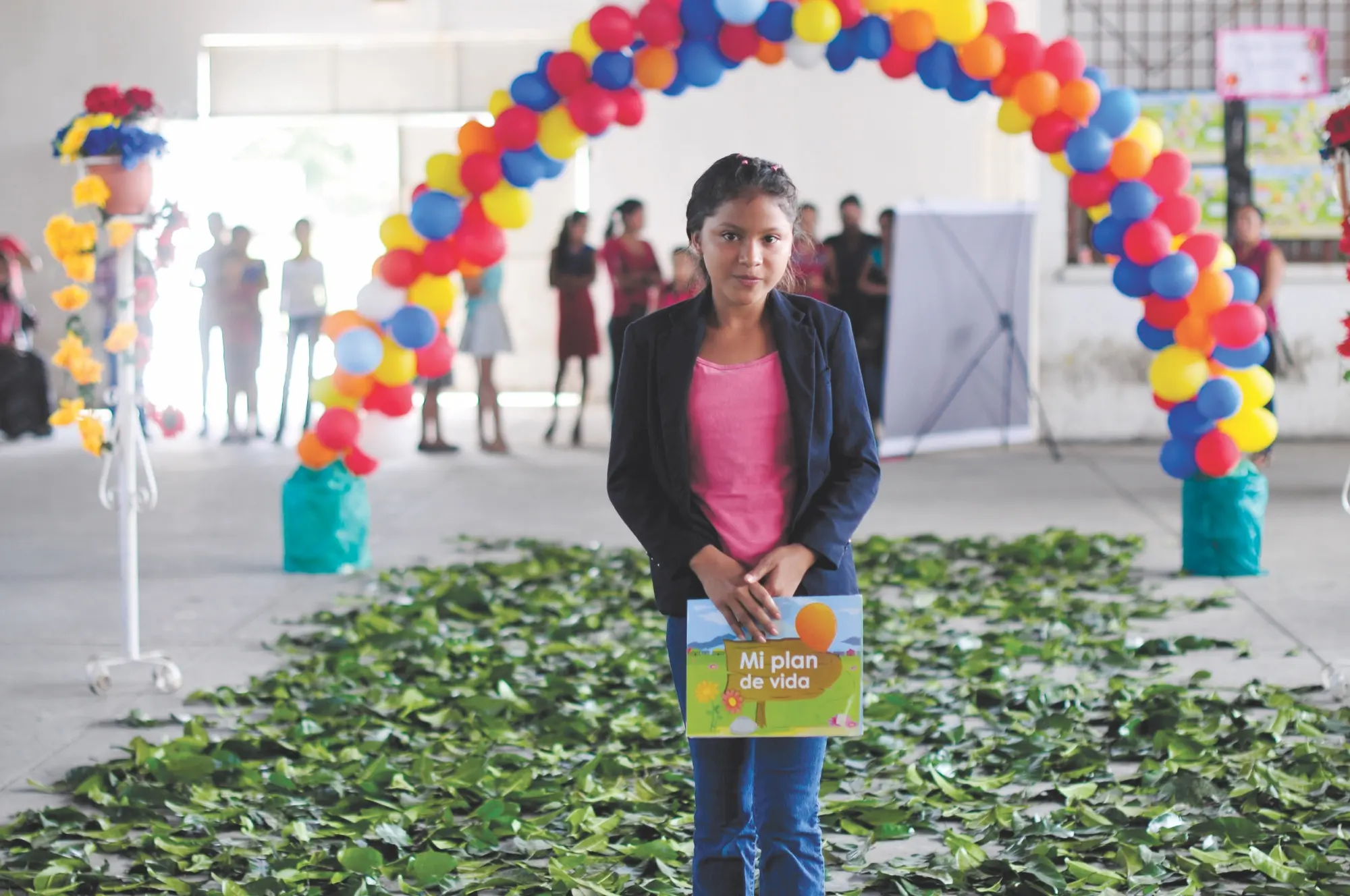 Una joven de pie sostiene un cuaderno titulado «Mi plan de vida» bajo un arco de globos de colores.