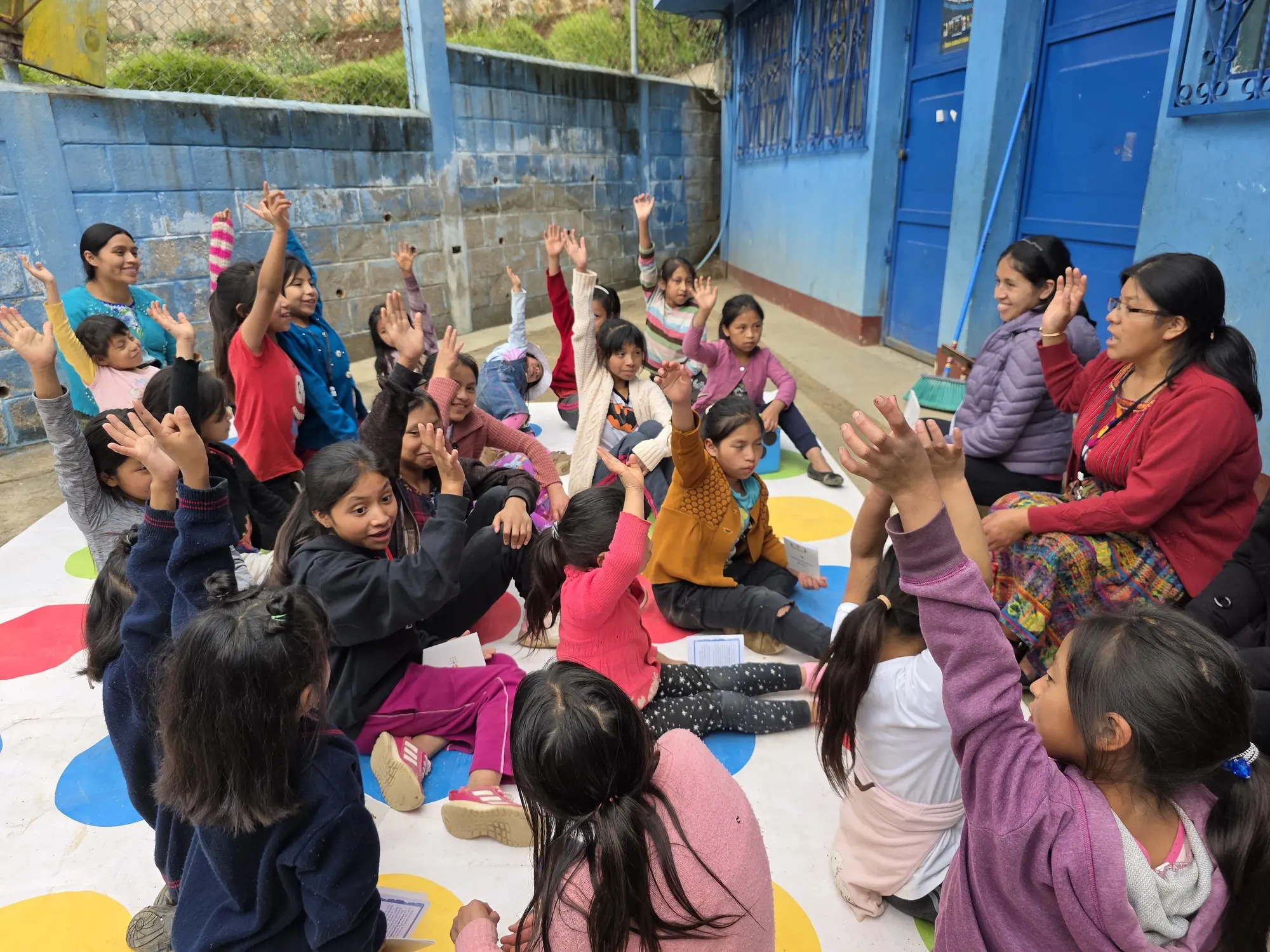Niñas y una mentora juegan sobre una lona de colores en el patio de una escuela.