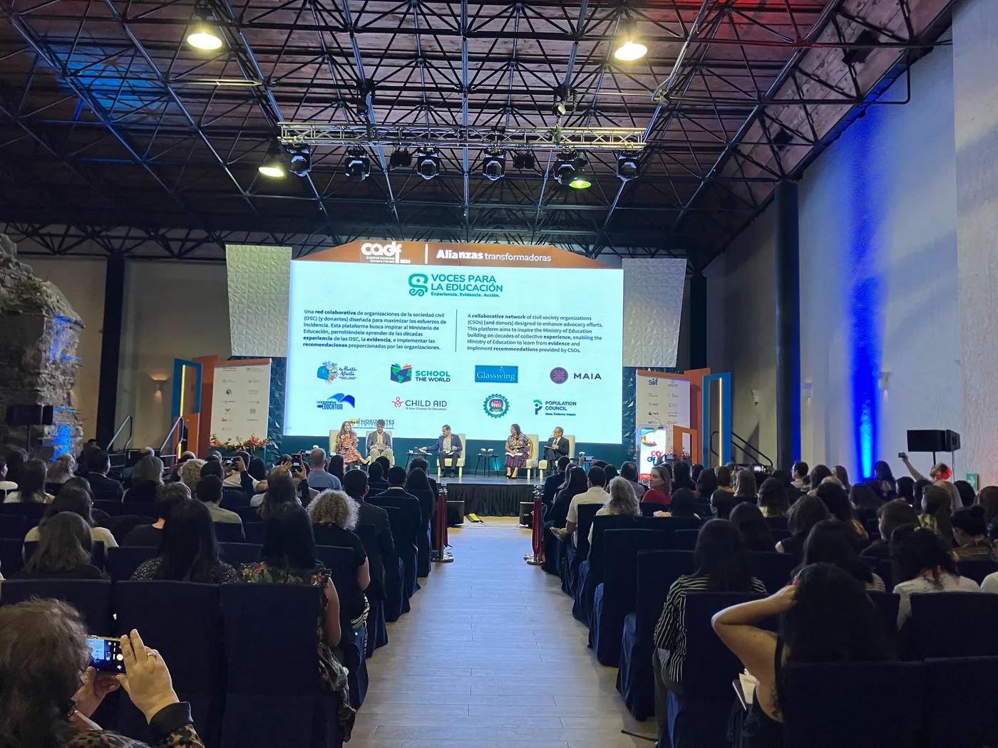 Un auditorio lleno sigue un panel sobre el escenario; en la pantalla, la presentación de Voces para la Educación con los logos de las organizaciones.