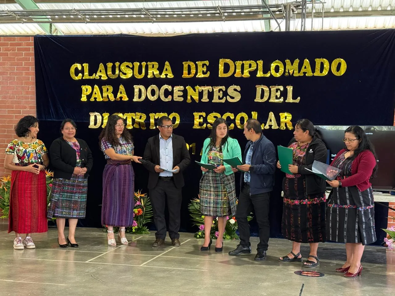 Ocho personas, varias con traje maya, leen documentos en el escenario frente a un telón que dice «Clausura de diplomado para docentes del distrito escolar».