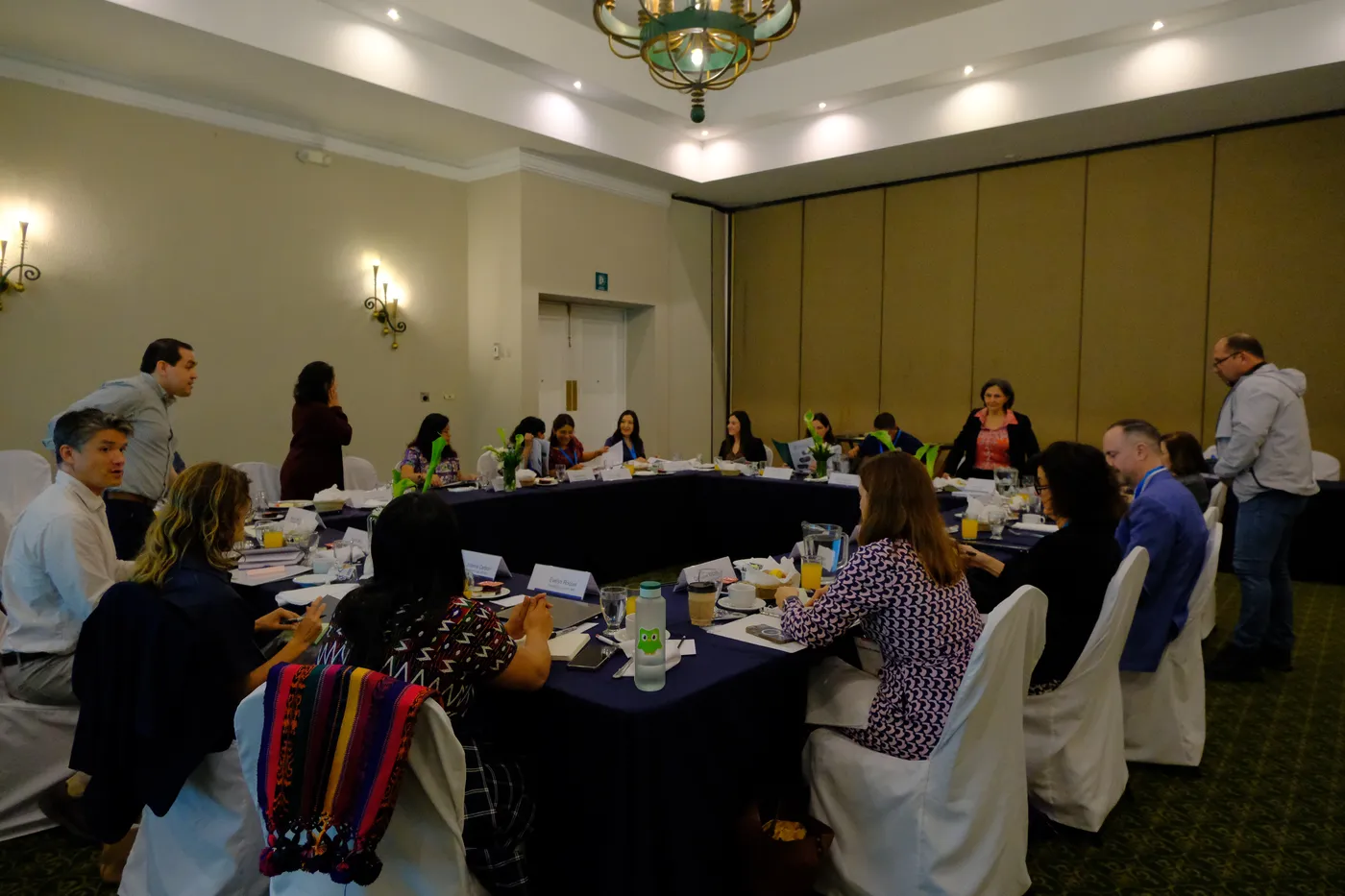 Personas conversan sentadas alrededor de mesas dispuestas en rectángulo en un salón.